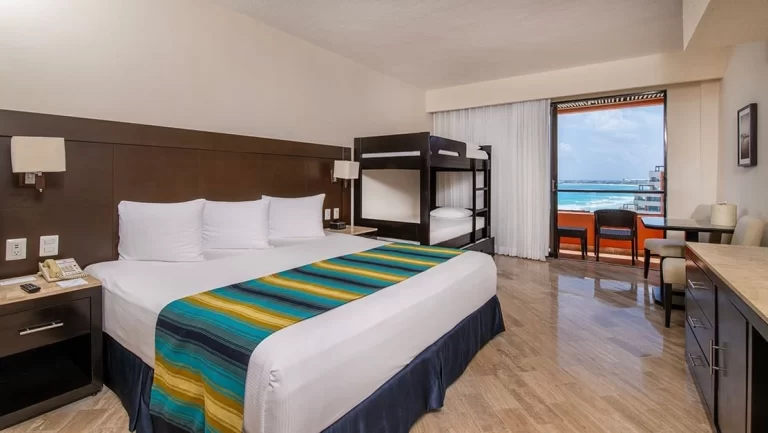 Habitación de hotel con cama king y litera, escritorio, balcón con vista al mar y decoración en tonos neutros.